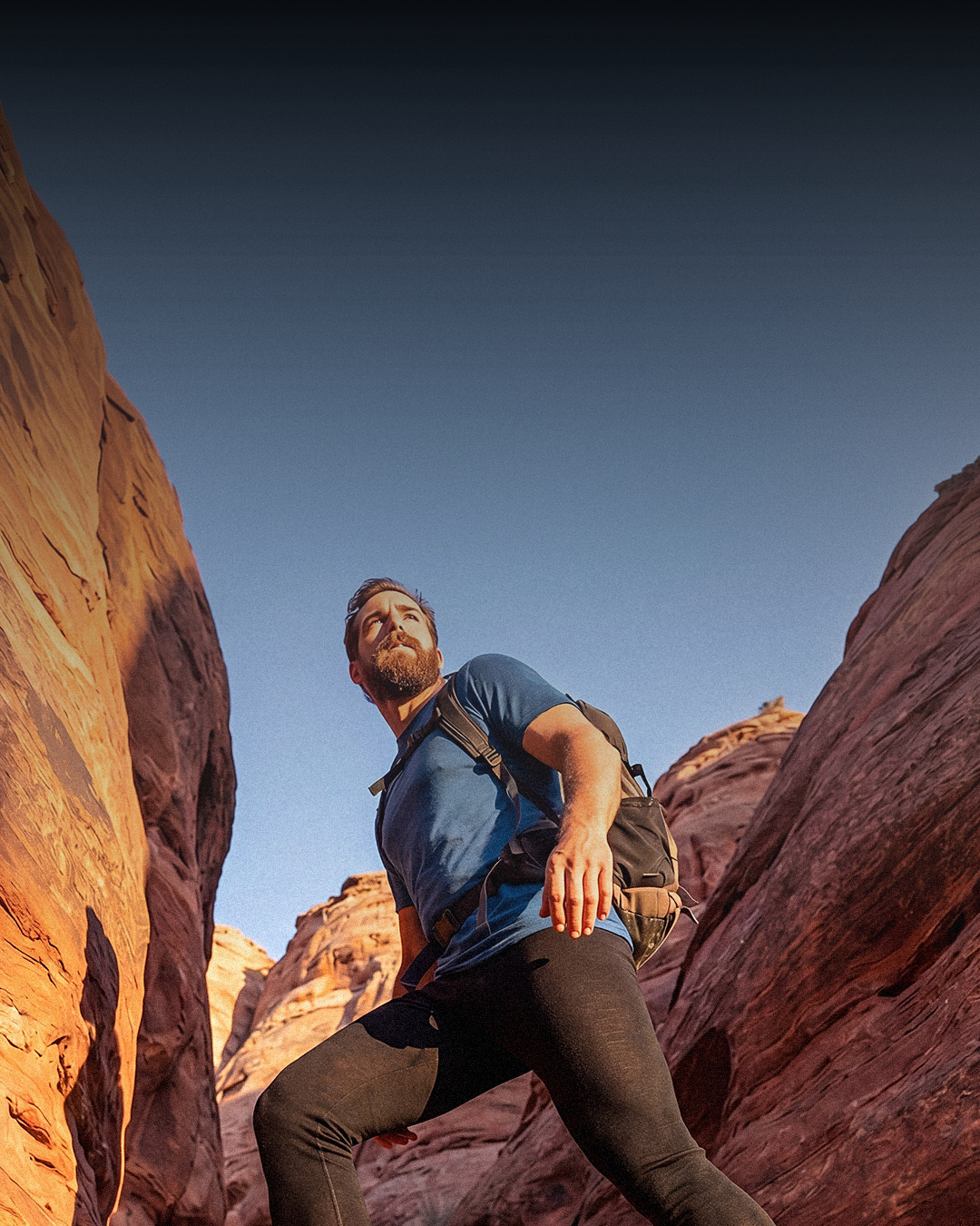 A low-angle outdoor lifestyle shot of a bearded man hiking up a rugged red rock canyon. He is captured mid-stride, stepping upward on the uneven terrain. He is wearing a short-sleeved blue t-shirt, black athletic leggings, sturdy brown hiking boots, and carrying a backpack. The warm, golden hour sunlight casts shadows and highlights the deeply striated texture of the canyon's rock walls. A narrow strip of clear blue sky is visible through the opening at the top left.