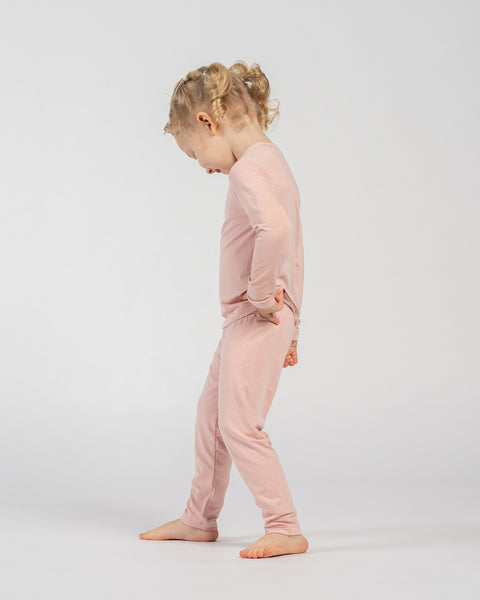 Child in pink bamboo long-sleeve top and matching pants, standing barefoot in a side profile pose with head slightly bent forward and hands resting on hips against a plain background.