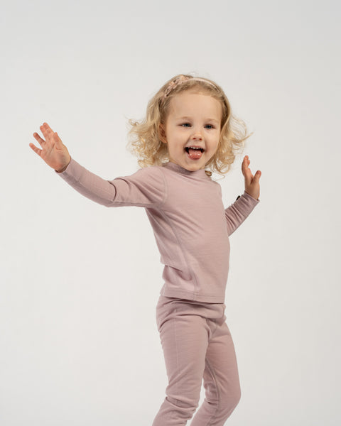 A playful child with blonde curly hair and a light pink floral headband stands against a white background, wearing a dusty pink long-sleeved top and matching pants. The child has their arms spread wide and their tongue slightly out, looking enthusiastically at the camera.