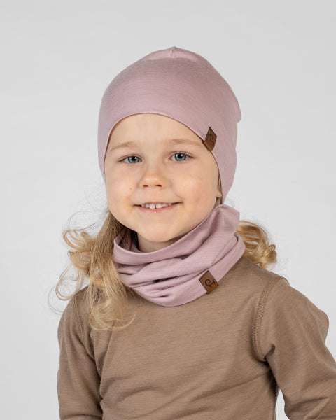 A studio portrait of a smiling young blonde girl looking directly at the camera showing her teeth. She is wearing a matching set of a dusty pink beanie hat and a dusty pink neck gaiter gathered comfortably around her neck. Both items feature a small brown leather logo patch. She is wearing a light brown long-sleeved shirt against a plain light grey background.