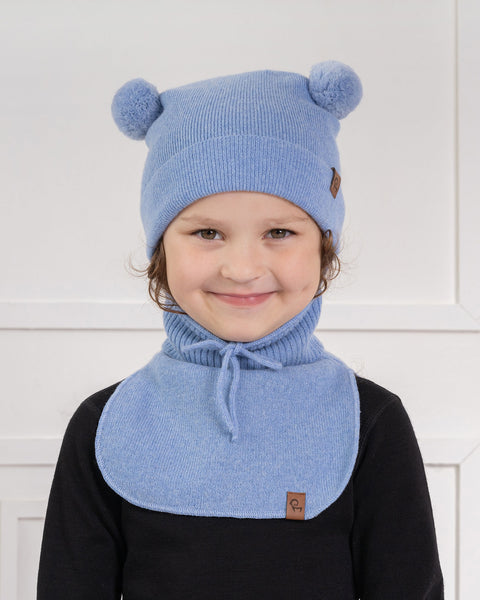 A young child with light brown curly hair and a subtle smile, is looking directly at the viewer. They are wearing a light blue knit hat with two light blue pom-poms on top and a small brown label on the side, and a matching light blue knit neck warmer with a tie, over a black long-sleeved shirt. The background is a white paneled wall.