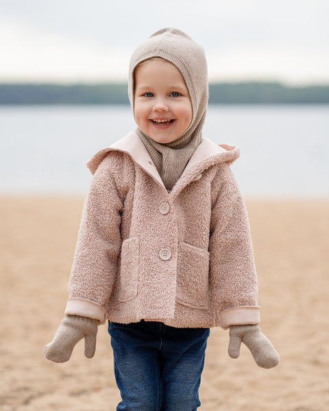 A happy young child with blonde hair and blue eyes, stands on a sandy beach. They are wearing a beige balaclava, a matching beige scarf, a light pink sherpa-style coat, beige mittens, and blue jeans, looking directly at the camera with a blurred body of water in the background.