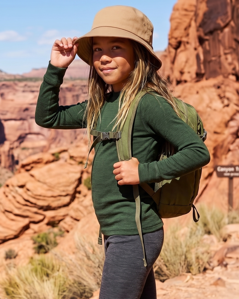 A child poses with one hand holding the brim of their tan bucket hat, standing outdoors with a rugged, red-rock canyon backdrop. They are wearing a dark green long-sleeve shirt, dark gray leggings, and a green backpack.