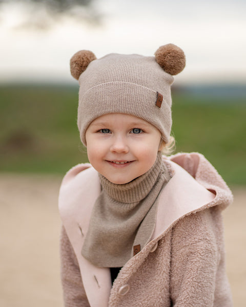 A young child with blue eyes and a slight smile, looks directly at the viewer. They are wearing a beige knit hat with brown pom-pom ears, a beige turtleneck or neck warmer, and a light pink sherpa-style coat. The background is a blurred outdoor scene with muted green and brown tones.