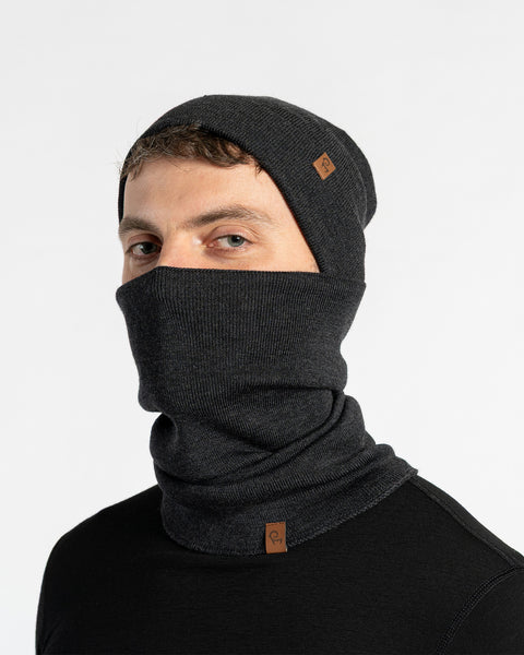 Man wearing dark gay knitted beanie and matching neck gaiter covering face, black top, studio background.