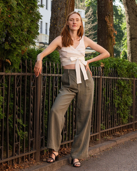 A fair-skinned woman with long reddish-brown hair stands outdoors, leaning against a dark metal fence. She is wearing a cropped menique wrap top that ties at the front and stone green wide-legged trousers, paired with black open-toe sandals. The background features lush green foliage, mature trees, and a glimpse of a light-colored building. The lighting suggests it is daytime.