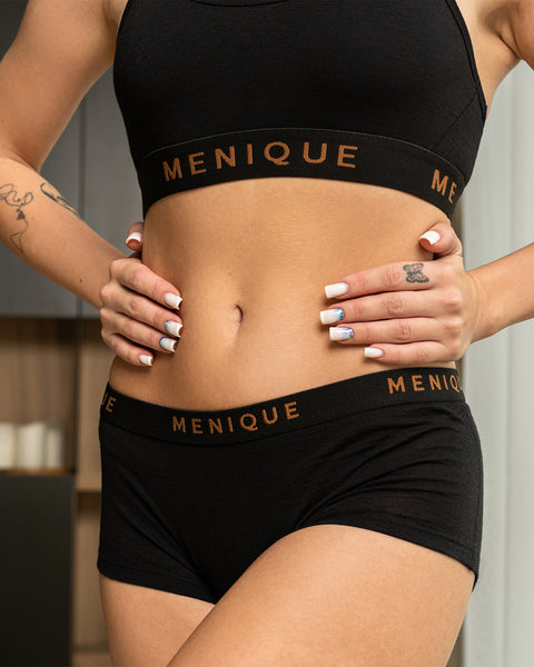 a woman wearing black women's merino boxer short underwear briefs, with her hands resting on her waist. She is wearing a matching bra as well.