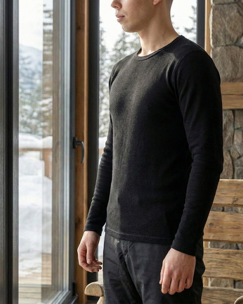 Man wearing black merino wool long sleeve top standing indoors by large window with snowy mountain view.
