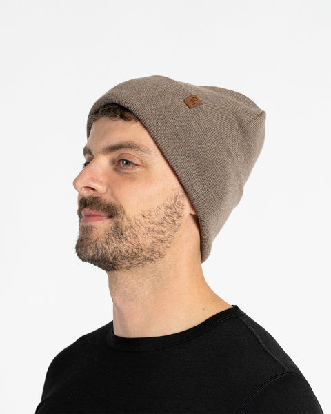 A person with light skin, short brown hair, and a beard is shown in profile against a white background. They are wearing a brown knit beanie with a small brown tag on the side. They are also wearing a black t-shirt.