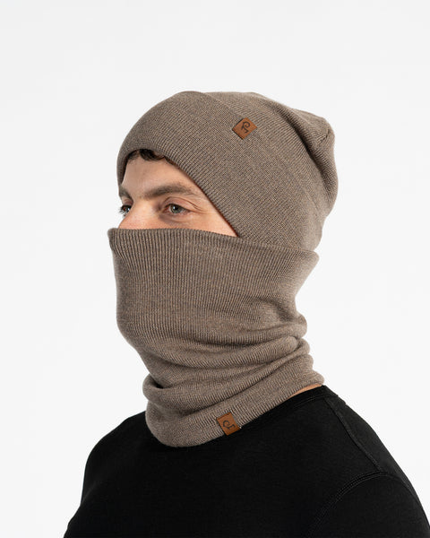 A male model, viewed in profile, wears a brown beanie and a matching neck gaiter that covers his nose and mouth. Both the beanie and gaiter feature a small, brown leather patch with a Menique logo. He has a short beard and dark hair. The background is solid white.