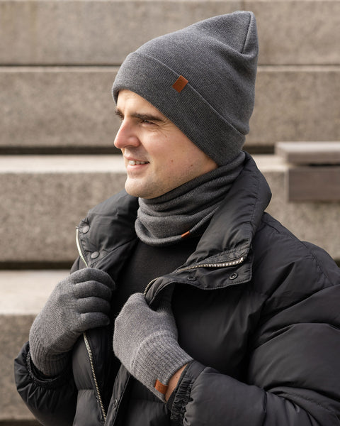 A smiling man is shown from the chest up, looking to the left. He is wearing a dark gray knitted beanie with a small brown leather tag on the front, a matching gray neck warmer, a black top, and a black puffer jacket. His hands, covered in gray knitted gloves, are clasped together near his chest. The background is a blurred outdoor setting with stone steps.