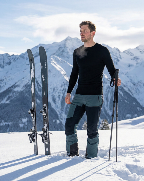 Man wearing a black merino wool base layer top and ski pants standing in deep snow with skis planted beside him, surrounded by snowy alpine mountains under a clear winter sky.