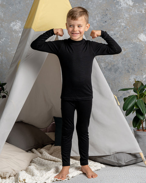 a boy wearing the menique Kids' 160 Thermal Long Sleeve Crew in Black color. He has his arms raised and bent, acting strong. He is barefoot on a carpet.