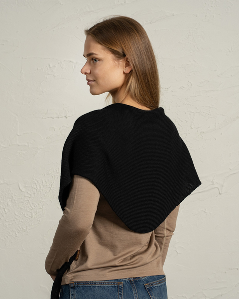 A woman standing sideways wears a black knitted merino bandana draped over her shoulders and upper back. She has straight, light brown hair and is dressed in a beige long-sleeve top and jeans, with a textured light wall in the background.