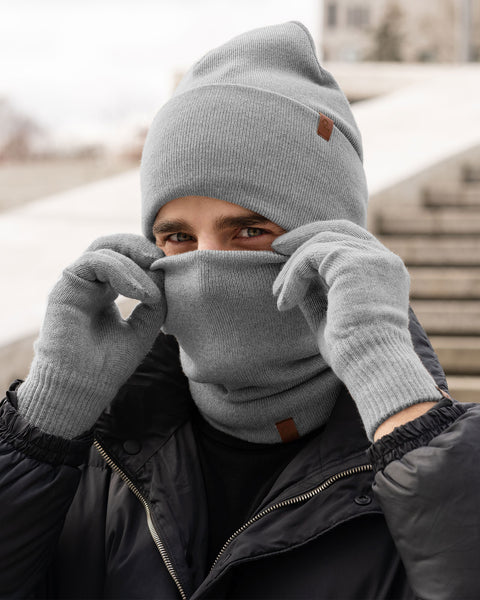 Man covering his face with a light gray Merino wool neck warmer while wearing matching beanie and gloves, styled with a black puffer jacket outdoors.