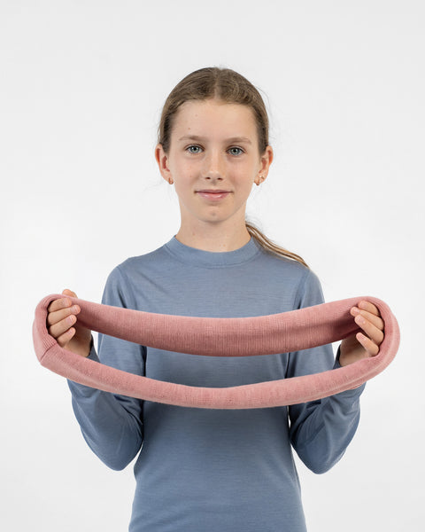 A young girl wearing a blue long-sleeved shirt, holding and stretching a pink knit infinity scarf horizontally in front of her.