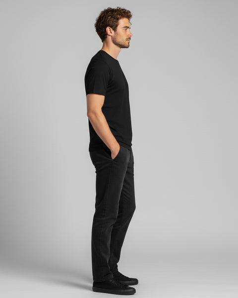 A man stands in profile wearing a black T-shirt, black jeans, and black sneakers against a light gray background, with hands in his pockets.