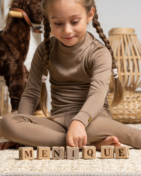 a girl wearing the menique Kids' Merino 160 Long Sleeve Crew in Beige color. She is playing with wooden letter cubes on the carpet.