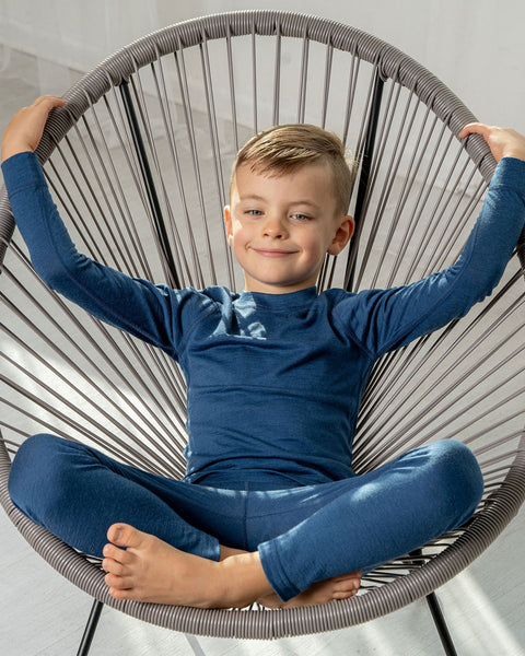 A boy sitting in a lounge chair, wearing the menique Kids' Merino 160 Long Sleeve Set in Denim color.