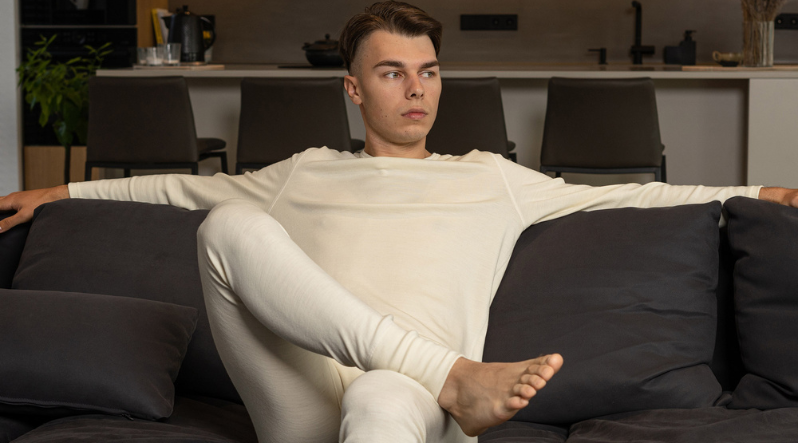 a man sitting on a couch, wearing the merino wool matching set in natural color, consisting of a long sleeve shirt and pants.