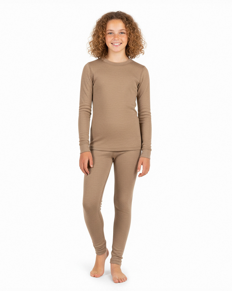 A girl wearing a beige long-sleeve Merino wool base layer top and matching leggings, standing barefoot and smiling against a white studio background.