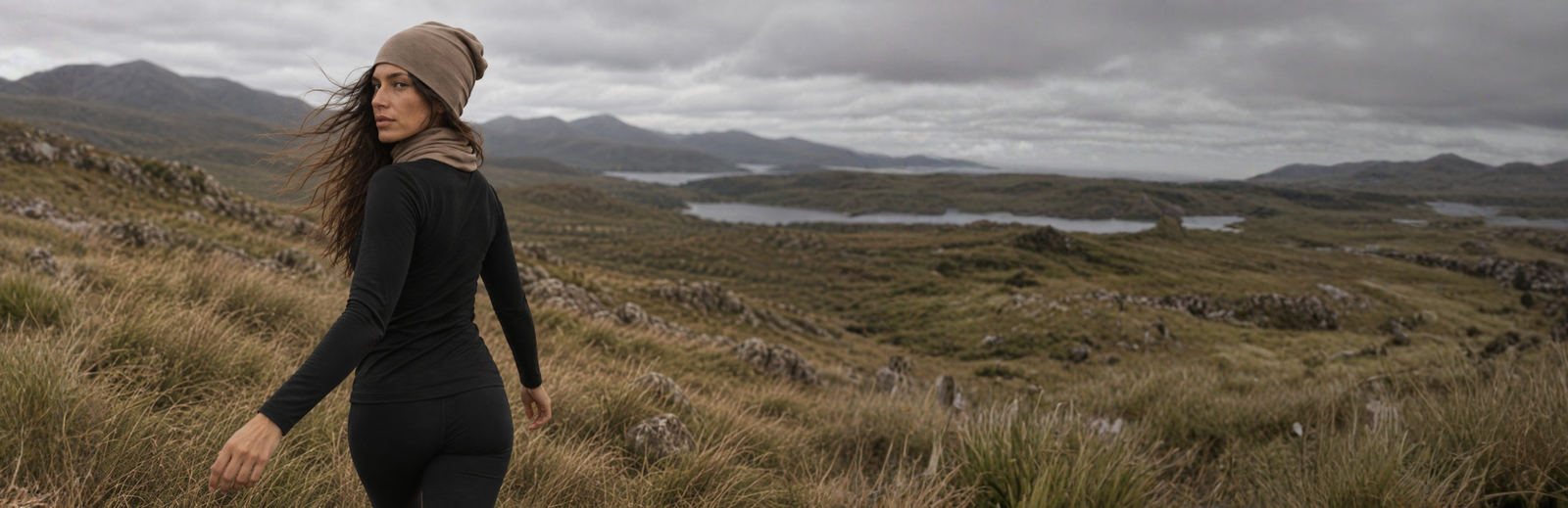 A woman with long wavy hair looks back over her shoulder while walking through a grassy mountain landscape. She is dressed in a black long-sleeved top and leggings, accessorized with a matching tan beanie and neck gaiter under a moody gray sky.