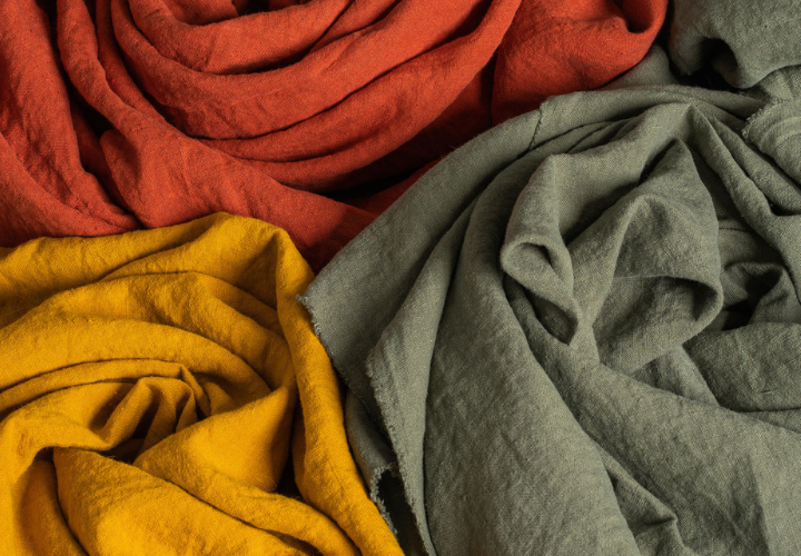Close-up of soft, crumpled linen fabric in warm earthy tones—rust red, mustard yellow, and sage green—showcasing the natural texture and richness of the material.
