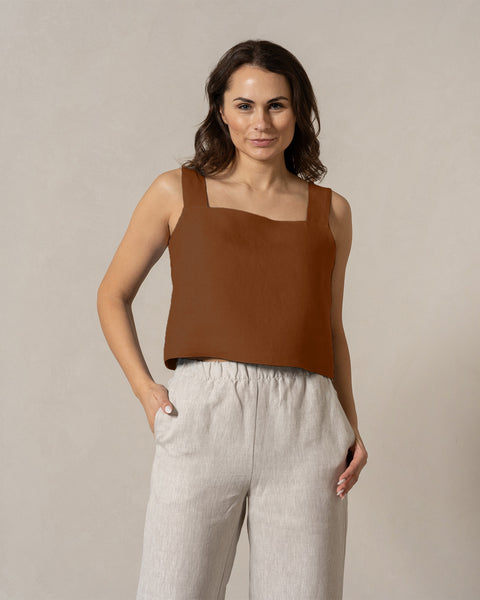 A woman with medium-toned skin and dark hair looks directly at the camera with a neutral expression. She is wearing a teal, sleeveless square-neck linen top and menique
 linen pants, with her one hand in the pocket. The background is a plain, light beige color.