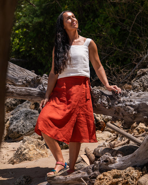 A woman wearing a white top and an cinnamon red skirt stands on a sandy area with rocks and driftwood, looking up with a smile.