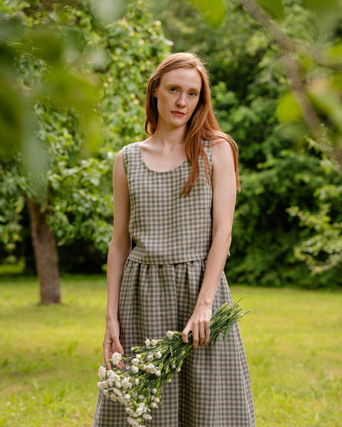 The model is wearing a two-piece set of the ANA crop top and SOPHIA skirt. She is holding a bouquet of flowers, slightly covering the front of the skirt. She is facing the camera, the background features a garden.