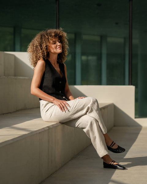 A woman with voluminous curly brown hair sits with her eyes closed on a set of concrete steps, seemingly enjoying the sunlight. She is wearing a dark menique sleeveless top, light-colored, loose-fitting linen trousers, and black ballet-style shoes with straps. The background features a modern building with large, dark windows, and the shadows suggest it is a bright day.