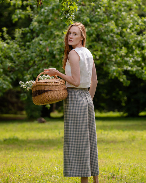 The model is posing sideways, revealing the elastic band at the back of her green gingham pattern linen pants. It is visible, that the pants are slightly cropped and a bit above her ankles. She is holding a basket full of flowers. 