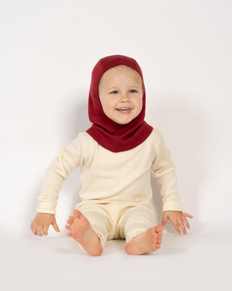 A toddler girl sitting on the ground, wearing the menique kids&#39; merino 160gsm balaclava in royal cherry color, studio background picture.