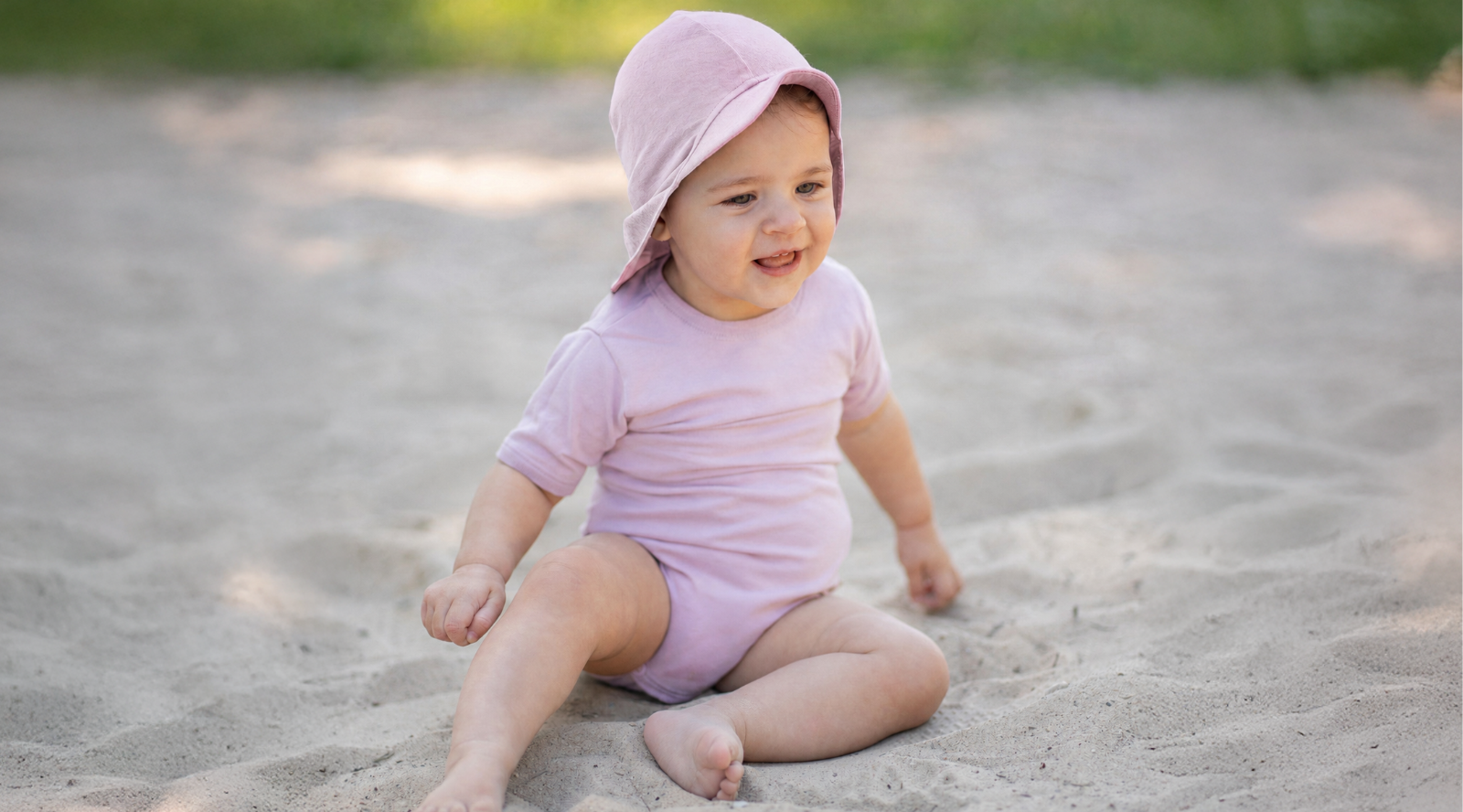 A smiling baby sitting barefoot in the sand outdoors, looking happily off to the side. The baby is wearing a light pink short-sleeve bodysuit and a matching dusty pink sun cap with a front brim and protective neck flap.