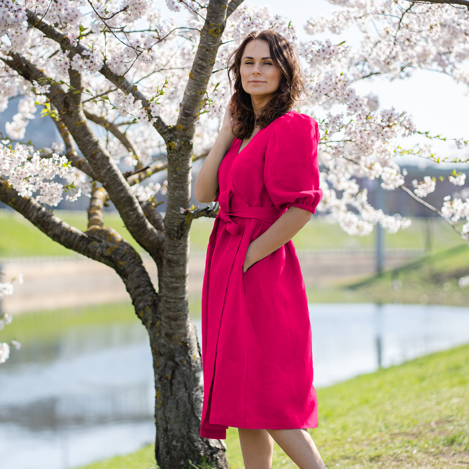 A woman with brown hair, wearing a mint green linen wrap dress with elbow-length puff sleeves, stands outdoors next to a tree with white blossoms.