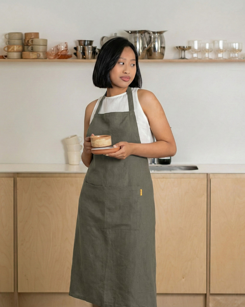 Woman wearing a stone green linen bib apron, standing in a modern kitchen and holding a ceramic cup and saucer.