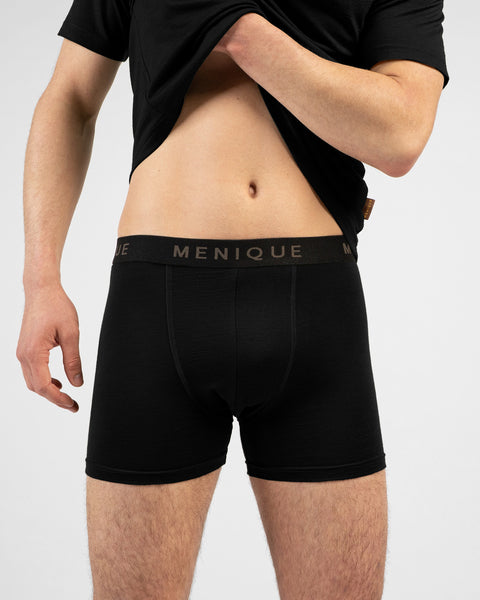 A close-up shot of a man standing against a white background, lifting his black t-shirt to reveal black boxer briefs with a branded "MENIQUE" waistband.