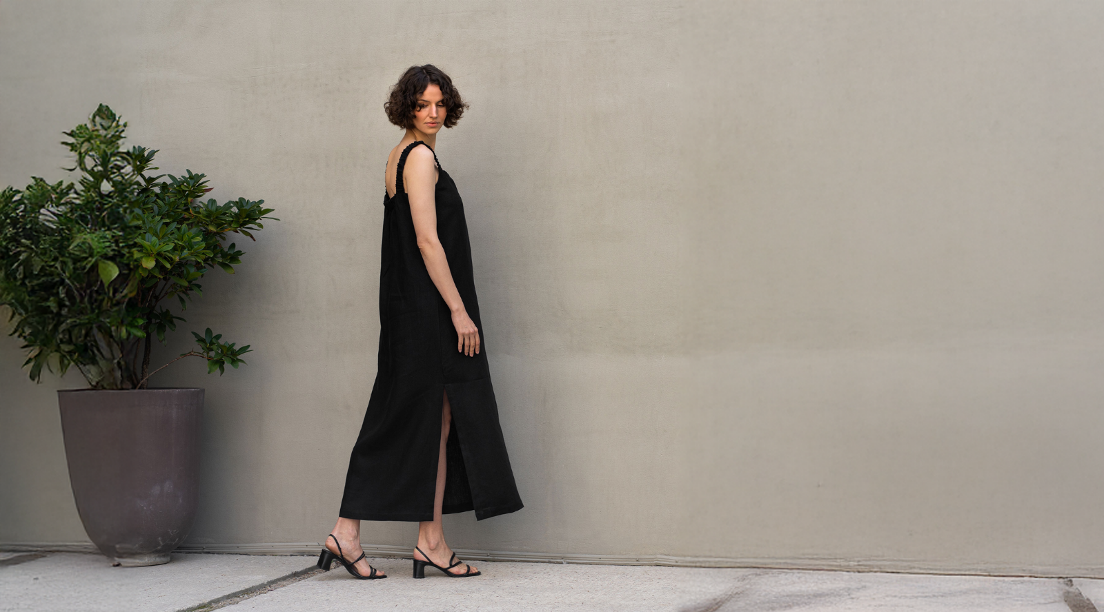 Woman wearing a minimalist black sleeveless midi dress with a side slit, standing against a neutral wall, styled with black heeled sandals for an elegant, modern look.