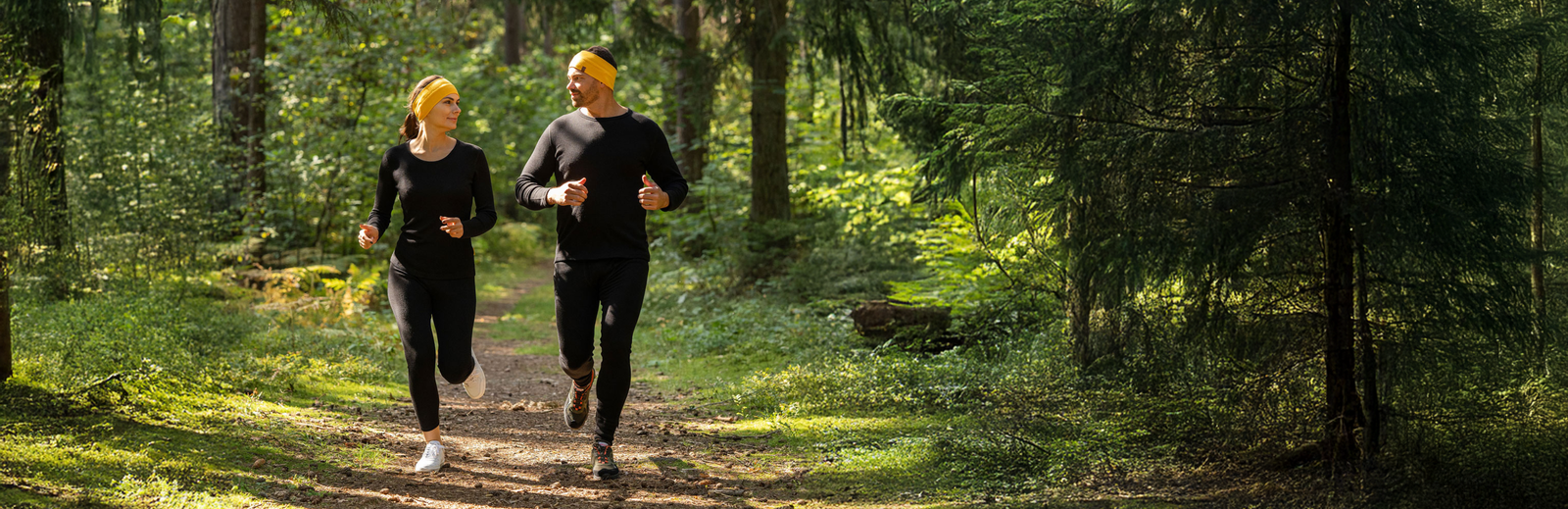 Man and woman running in forest wearing merino wool activewear and headbands, breathable moisture-wicking clothing for outdoor sports.