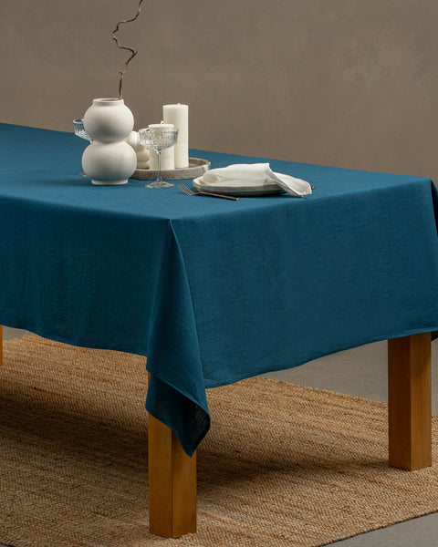 The menique linen tablecloth in cobalt blue color, with a modern white vase on top of it and two different height white candles by the sides.