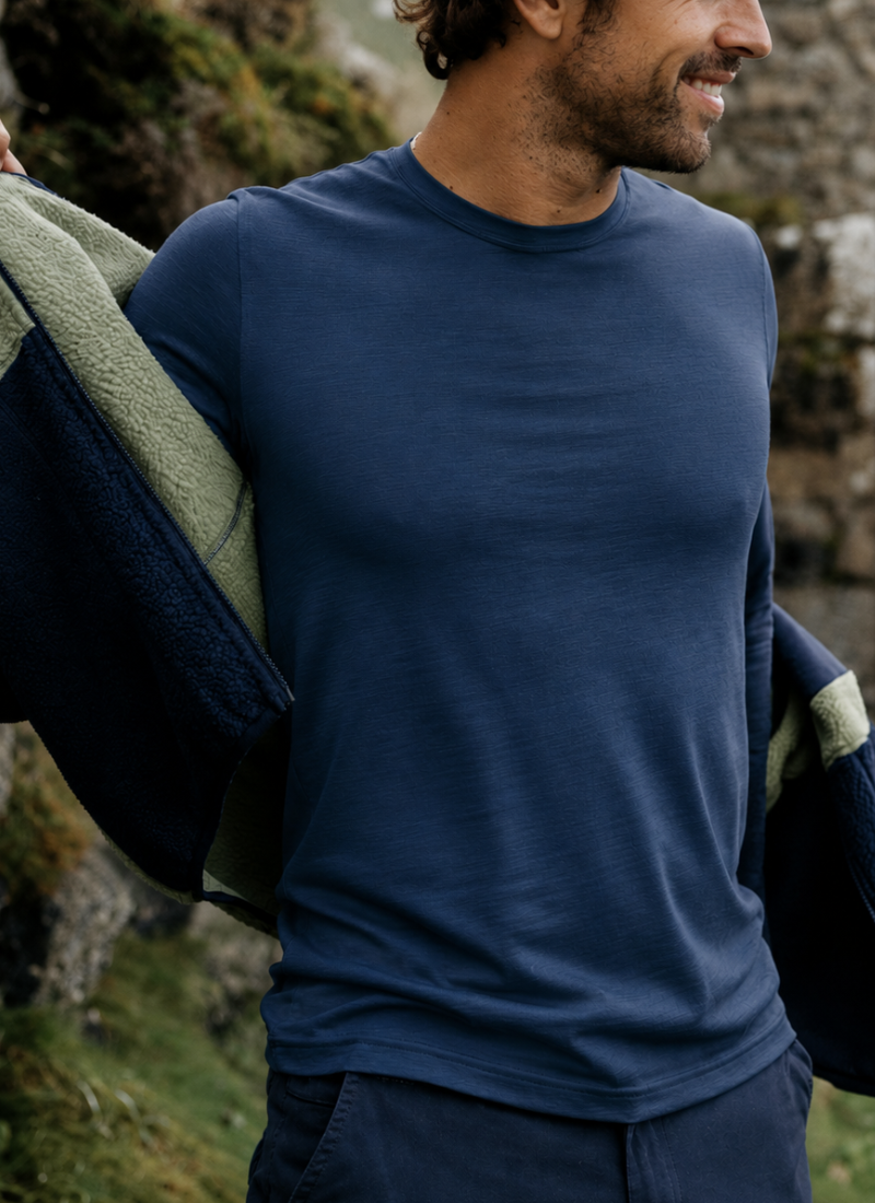 A man wearing a navy blue crew-neck long-sleeve shirt, with a dark navy and light green fleece jacket draped over his shoulders, set against a natural outdoor background of rocks and grass.