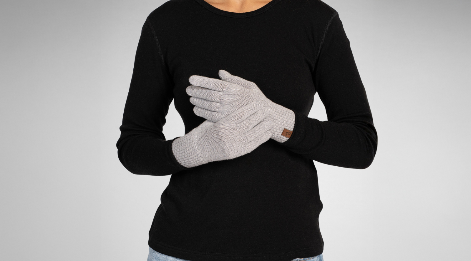 A person wearing a black long-sleeved shirt crosses their hands in front of their torso. They are modeling a pair of light grey knitted gloves that feature a small brown logo tag on the cuff.