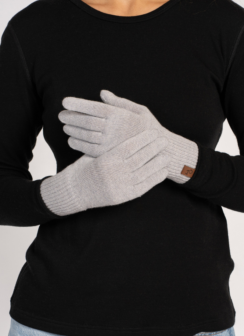 A person wearing a black long-sleeved shirt crosses their hands in front of their torso. They are modeling a pair of light grey knitted gloves that feature a small brown logo tag on the cuff.