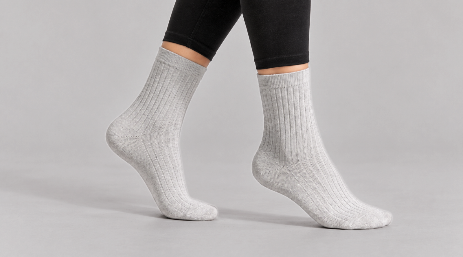 The cropped image focuses on a person's lower legs and feet as they take a mid-air step against a plain light grey studio background. They are wearing black leggings seamlessly paired with snug, light grey ribbed crew socks.