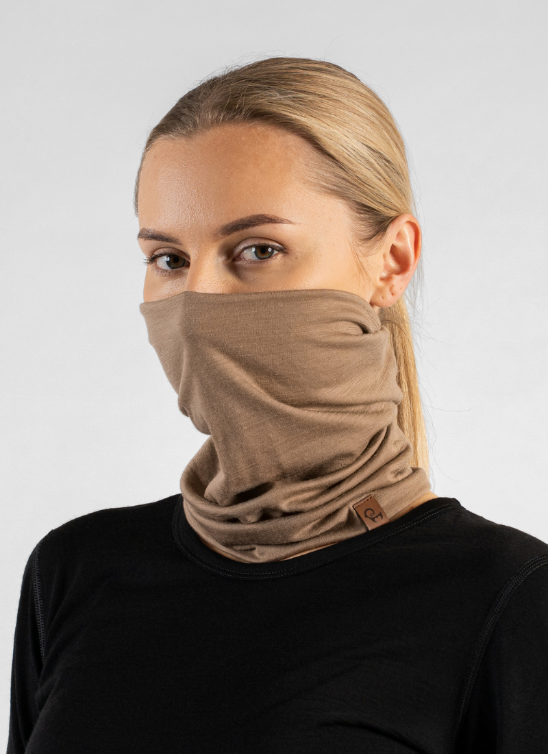 A blonde woman with her hair pulled back looks directly at the camera against a seamless light grey studio background. She is modeling a tan knit neck gaiter pulled up over her nose and mouth, paired with a black long-sleeved top.