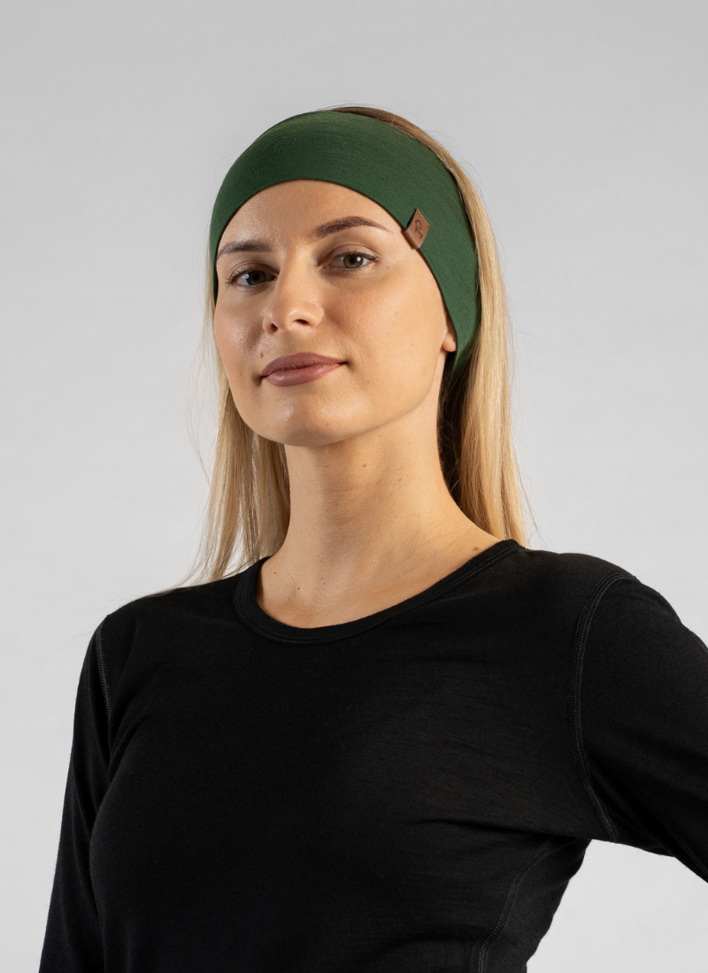 A blonde woman stands against a light grey studio backdrop modeling a wide, dark green knit headband. She pairs the accessory with a simple black long-sleeved top while looking directly toward the camera with a neutral expression.