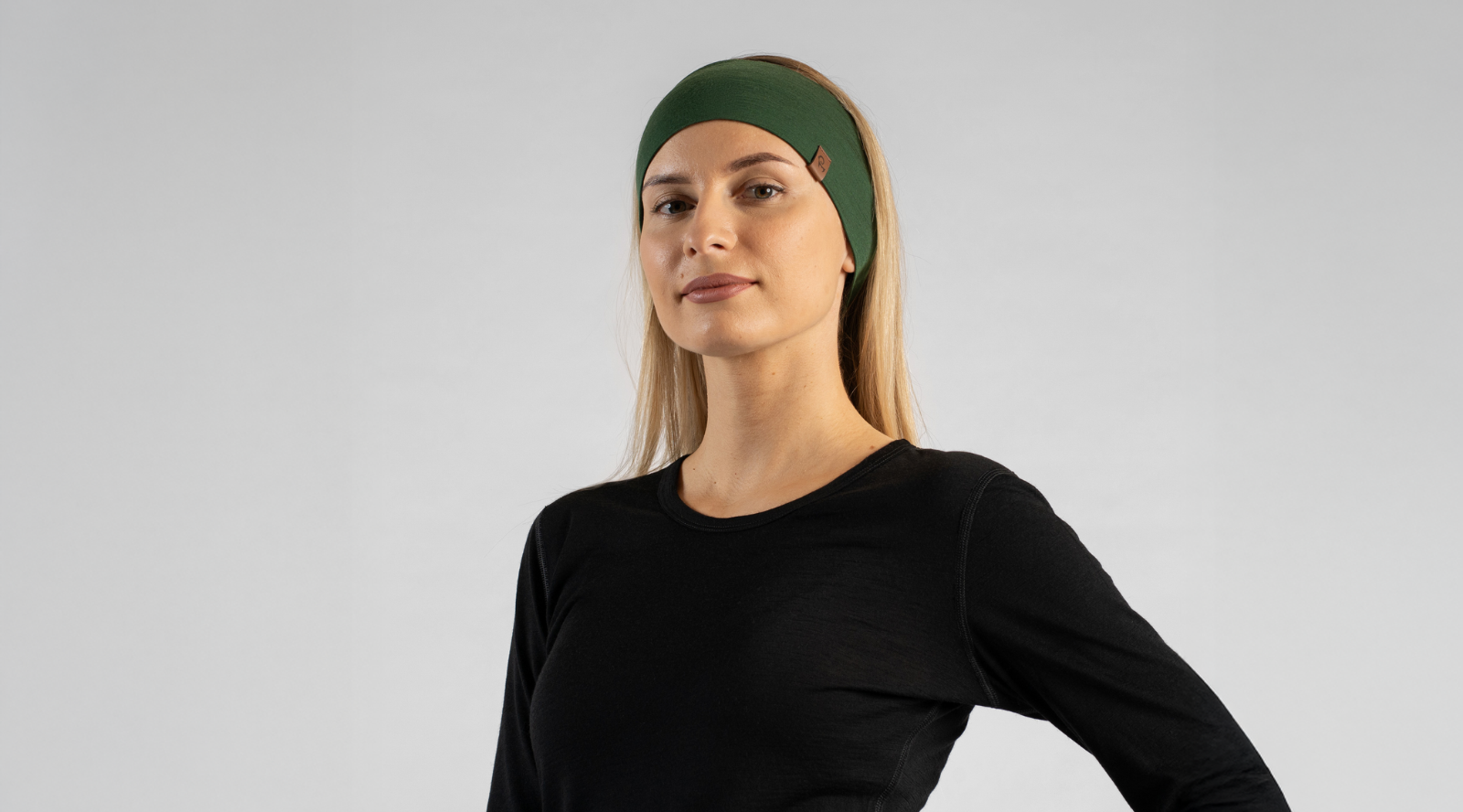 A blonde woman stands against a light grey studio backdrop modeling a wide, dark green knit headband. She pairs the accessory with a simple black long-sleeved top while looking directly toward the camera with a neutral expression.