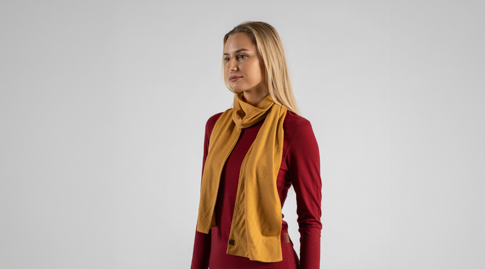 A blonde woman stands against a seamless light grey studio background, looking slightly off-camera with a neutral expression. She is wearing a dark red long-sleeved top with a mustard yellow knit scarf draped casually around her neck.