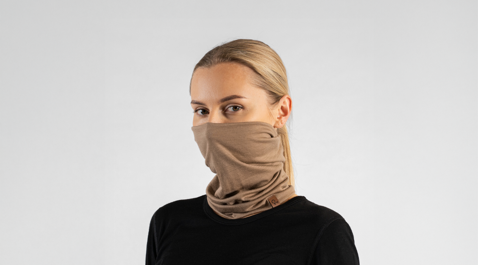 A blonde woman with her hair pulled back looks directly at the camera against a seamless light grey studio background. She is modeling a tan knit neck gaiter pulled up over her nose and mouth, paired with a black long-sleeved top.