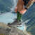 Close-up of a hiker wearing green ribbed Merino hiking socks with brown hiking boots, standing on a rock with a mountain lake in the background.